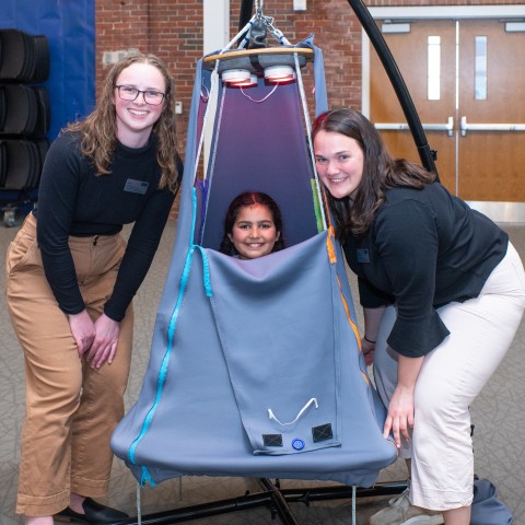 A child sits inside a suspended tent and two students pose around her